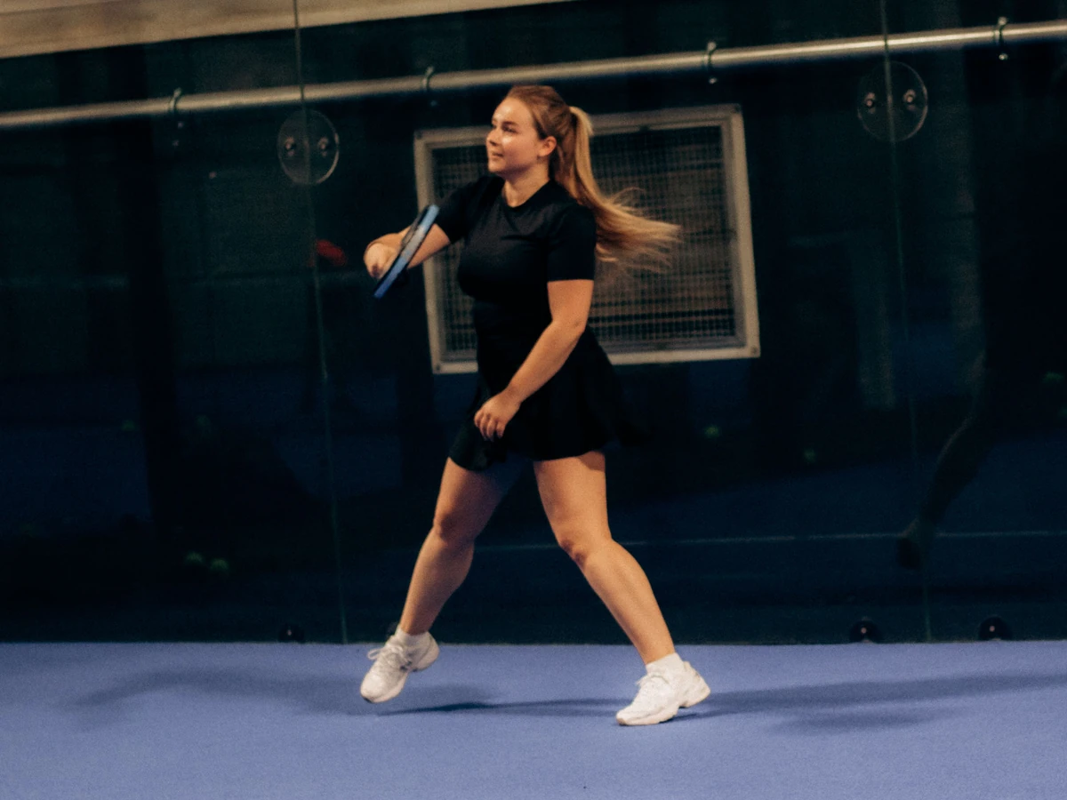 A woman practises a gentle swing on a blue indoor padel court.