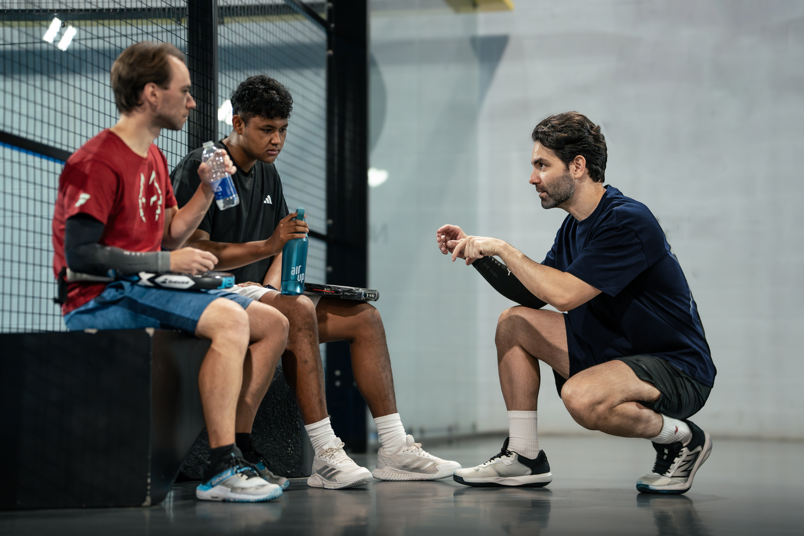 A coach crouches to talk with two players sitting beside the court.