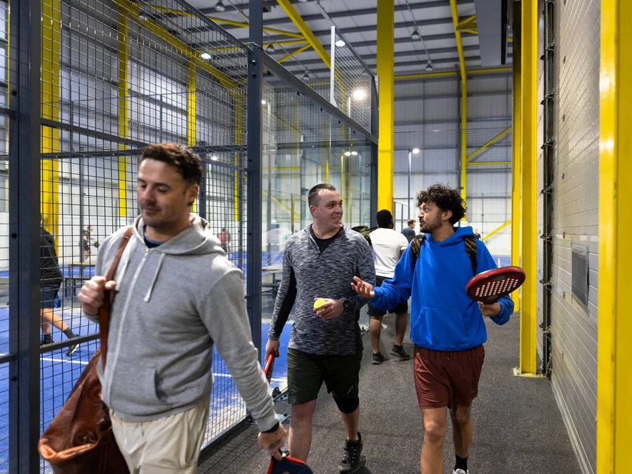 Three players arriving at indoor padel courts with rackets and sports bags.
