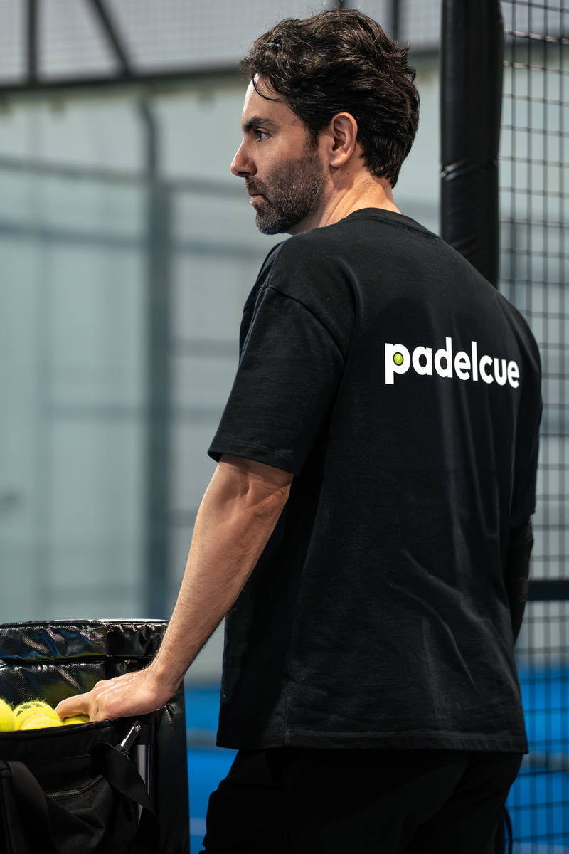 A coach stands beside a basket of padel balls on an indoor padel court.