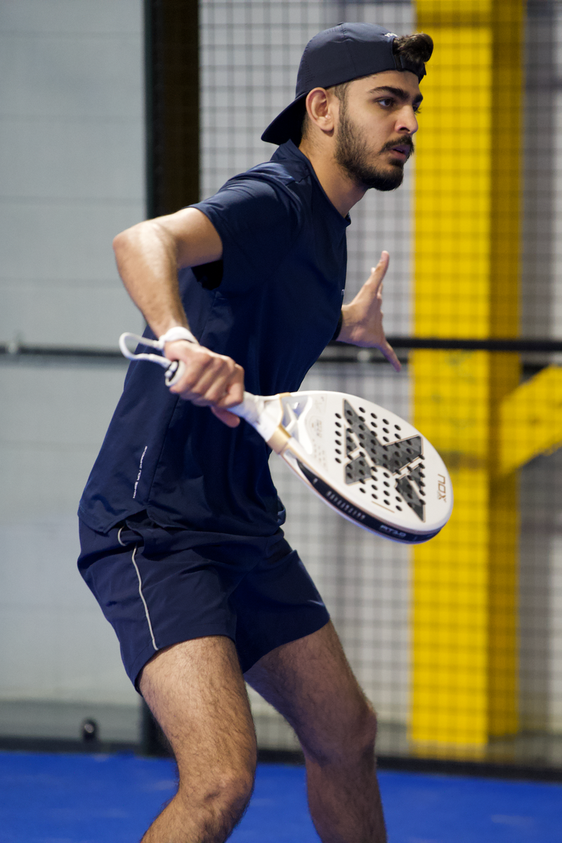 A player in navy kit and a backwards cap prepares a forehand on an indoor padel court.